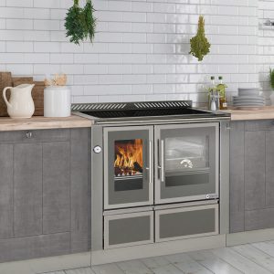 Küchenzeile mit grauen Schränken, Holzarbeitsplatte, Holzofen mit Sichtfenster und Fliesenspiegel aus weißen rechteckigen Fliesen.