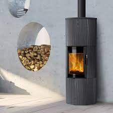 Moderner grauer Holzofen mit sichtbarer Flamme, daneben Holzstapel in Wandnische, darüber Wandregal mit Büchern und Dekoration.