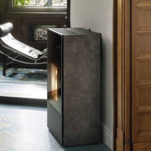 Moderner, grauer Kaminofen mit Sichtfenster steht an weißer Wand neben Holztür, im Hintergrund Ledersessel vor Fenster