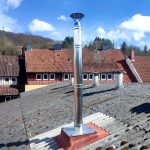 Metallkamin mit Abdeckung auf Schornstein mit rotem Ziegelmauerwerk auf Dach mit grauen Dachziegeln, im Hintergrund Häuser und bewölkter Himmel