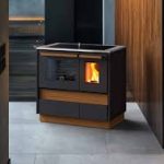 Kompakter Holzofen mit Glastür und sichtbarer Flamme, umgeben von dunklem Holz und grauem Boden.