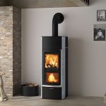 Moderner Kaminofen mit sichtbarem Feuer, schwarzer Front und grauen Seiten, an grauer Wand neben Steinmauer und Holzkorb