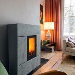 Moderner grauer Kamin mit sichtbarem Feuer, daneben ein Beistelltisch mit Lampe und Vorhang vor einem Fenster.